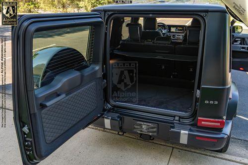 A black, armored Mercedes G63 SUV with its rear door open, revealing a spacious cargo area and the back seats. The vehicle is parked on a paved surface near a grassy area.