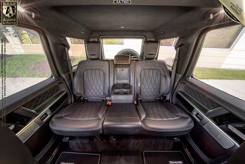 The image shows the interior of an Alpine Armoring armored Mercedes-Benz G63 vehicle with black leather seats, including a central fold-down armrest, against a backdrop of a building and greenery outside.