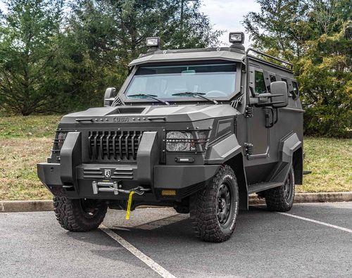 A heavily armored SWAT APC CUDA with off-road tires parked on an asphalt surface, with trees in the background.