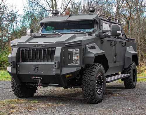 A heavily armored black Pit-Bull VXT is parked outdoors on a gravel surface with trees in the background.