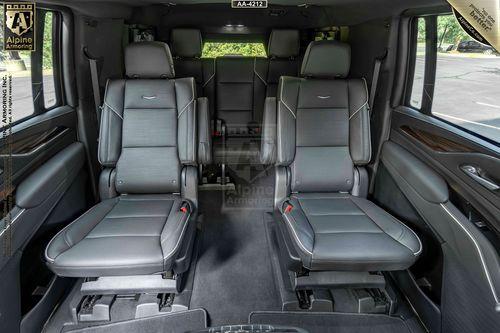 Interior view of a bulletproof Cadillac Escalade ESV SUV showing four leather seats, with two in front and two in the rear, separated by an aisle. The seats are black with detailed stitching, and the windows reveal a green outdoor scene.