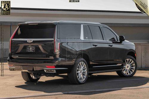 A black bulletproof Cadillac Escalade ESV SUV is parked on a paved surface near a concrete barrier. It features tinted windows and chrome details, with a badge reading "Escalade" on the rear. Logos are visible at the top corners of the image.