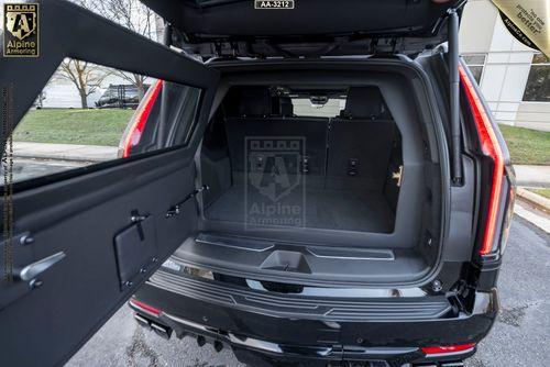 An empty Cadillac Escalade ESV SUV trunk with open tailgate showing a spacious cargo area. The vehicle is parked outside with grass and a building in the background. A badge showing "Alpine Armoring" is visible.